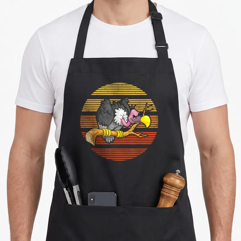 Funny vulture bird apron - Cartoon grey vulture on brown branch with yellow orange striped sun background — shown on a black cotton canvas apron against a white background