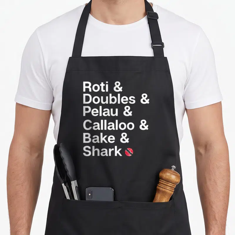 Trinidad food aprons - White text listing Roti, Doubles, Pelau, Callaloo, Bake, Shark with small circular red emblem — shown on a black cotton canvas apron against a white background