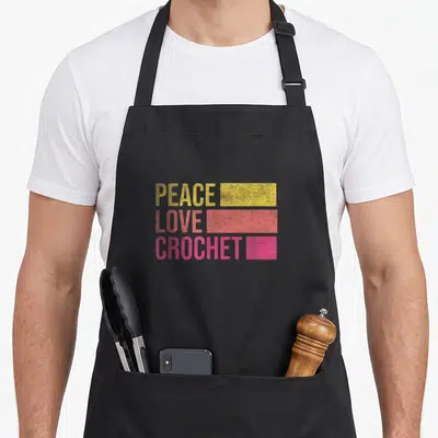 Funny crocheting crochet apron - PEACE LOVE CROCHET text with yellow, coral, and pink rectangular bars — shown on a black cotton canvas apron against a white background