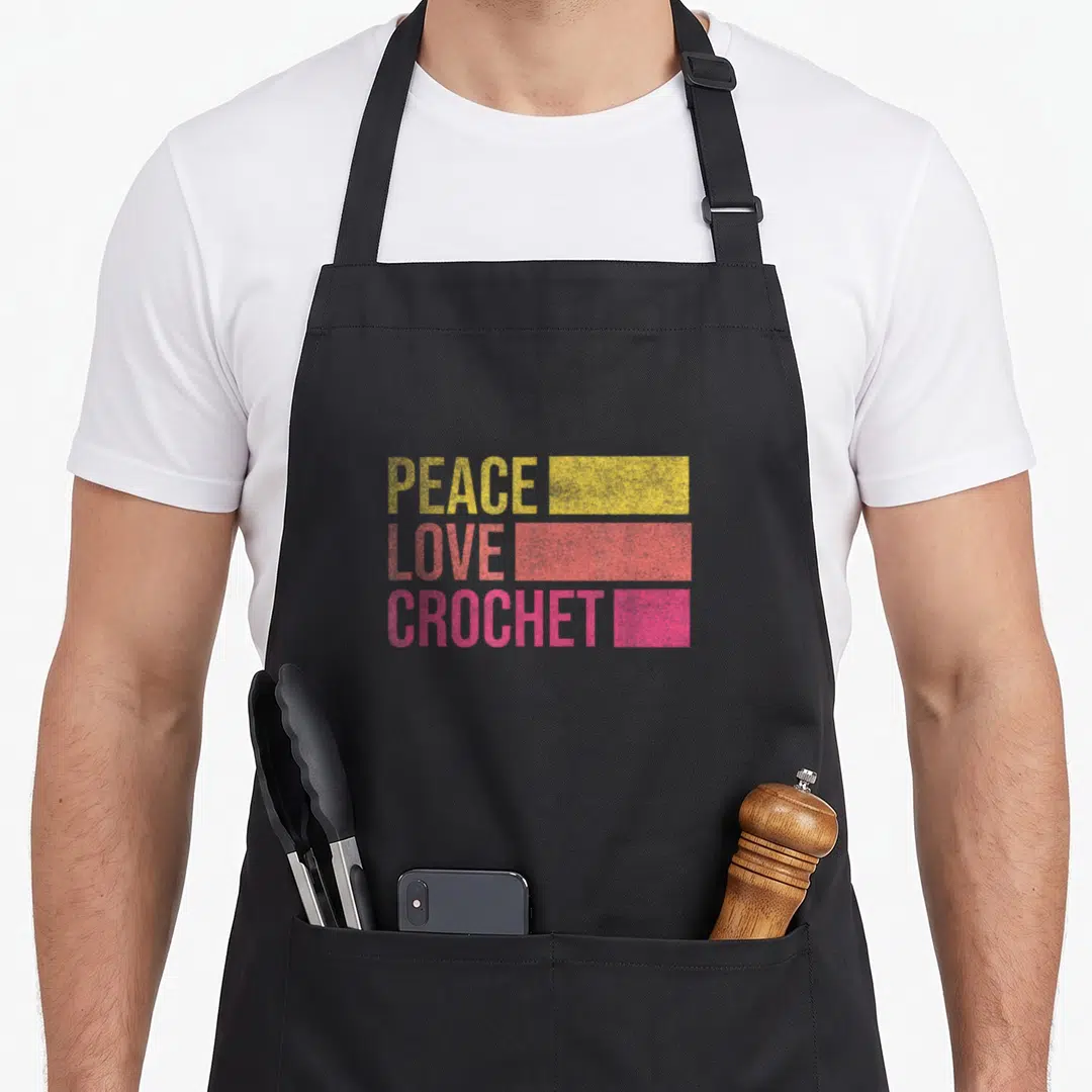 Funny crocheting crochet apron - PEACE LOVE CROCHET text with yellow, coral, and pink rectangular bars — shown on a black cotton canvas apron against a white background