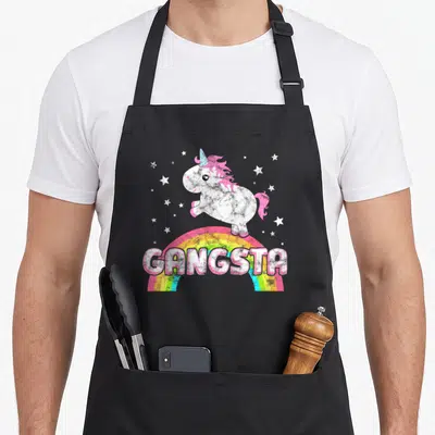 Ironic unicorn apron - White cartoon unicorn on rainbow arch with GANGSTA text and white stars — shown on a black cotton canvas apron against a white background
