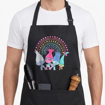 Funny troll rainbow apron - Four cartoon trolls under a rainbow arch of pink stars, blue dots, yellow triangles — shown on a black cotton canvas apron against a white background