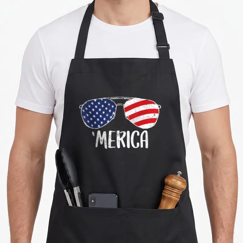 Sunglass 4th of july apron - Sunglasses with US flag lenses and MERICA text in distressed style — shown on a black cotton canvas apron against a white background
