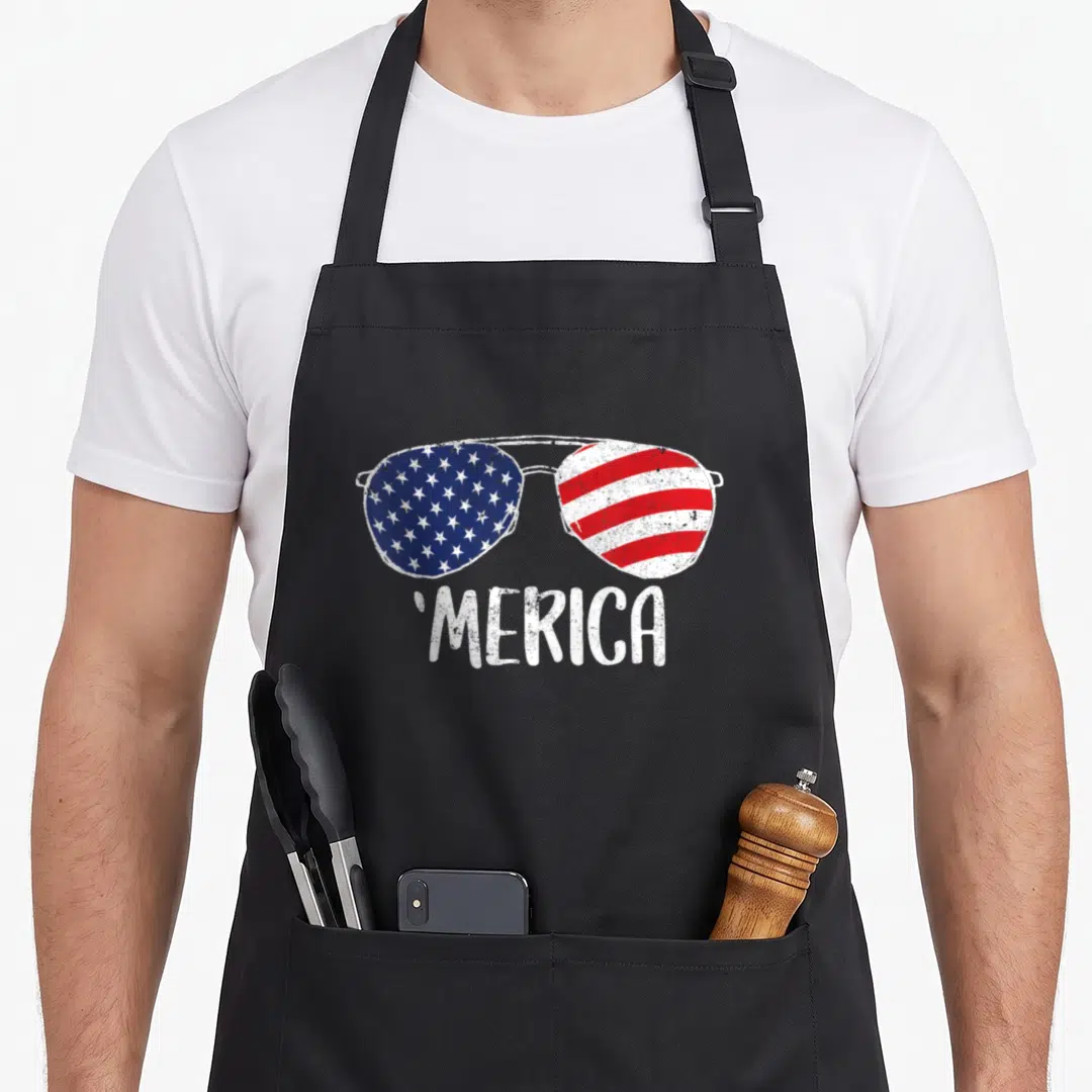 Sunglass 4th of july apron - Sunglasses with US flag lenses and MERICA text in distressed style — shown on a black cotton canvas apron against a white background
