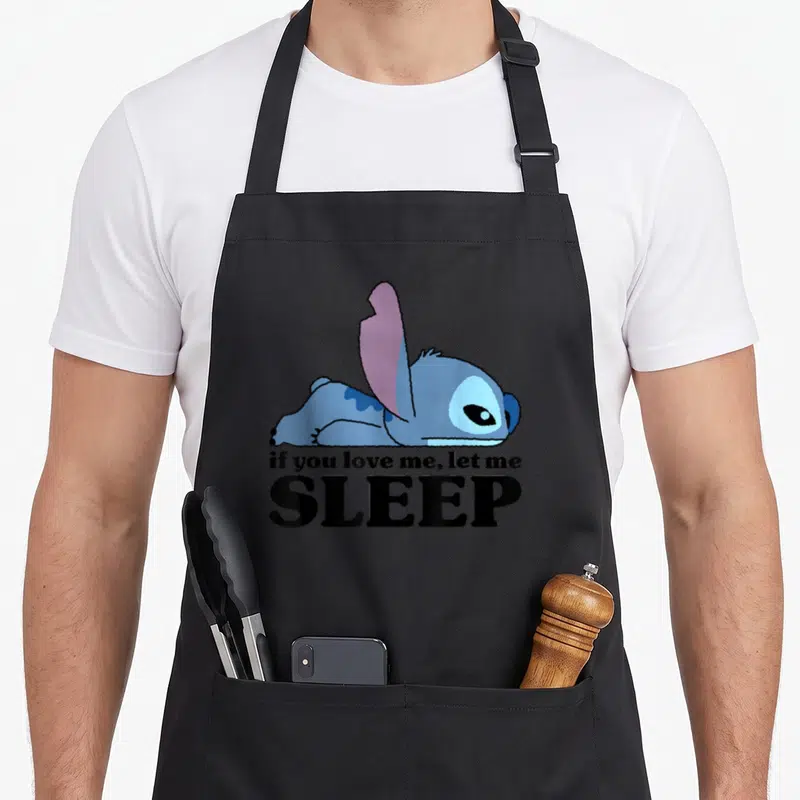 Sleepy stitch apron - Blue cartoon character lying flat with text if you love me let me SLEEP — shown on a black cotton canvas apron against a white background