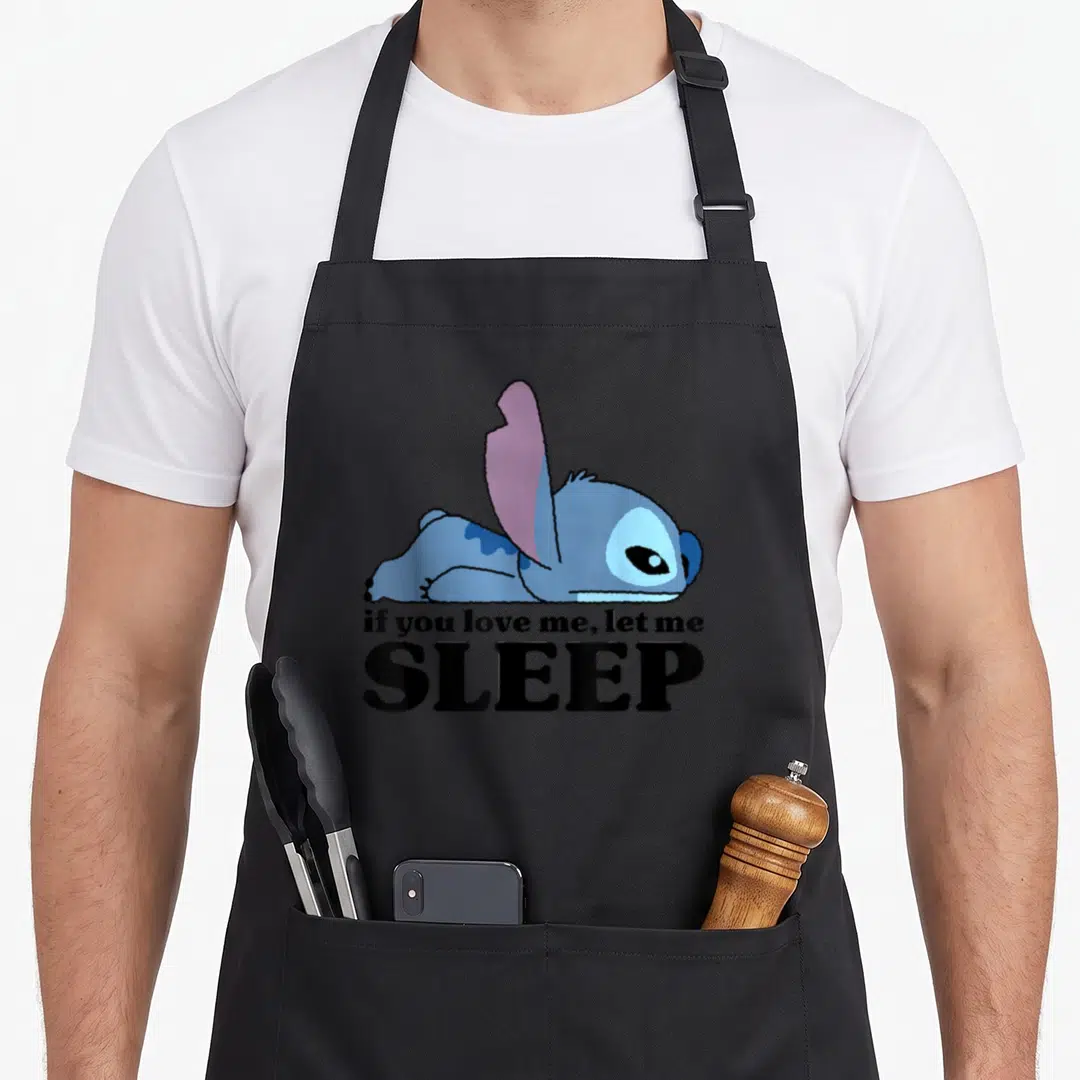 Sleepy stitch apron - Blue cartoon character lying flat with text if you love me let me SLEEP — shown on a black cotton canvas apron against a white background