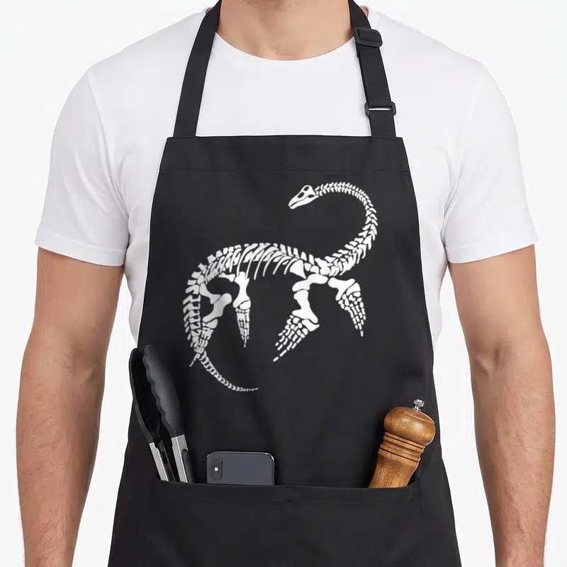 Plesiosaurus science apron - white plesiosaurus skeleton illustration on black background — shown on a black cotton canvas apron against a white background