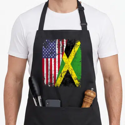 Jamaican root american flag apron - Two overlapping distressed flags in red white blue green yellow and black — shown on a black cotton canvas apron against a white background