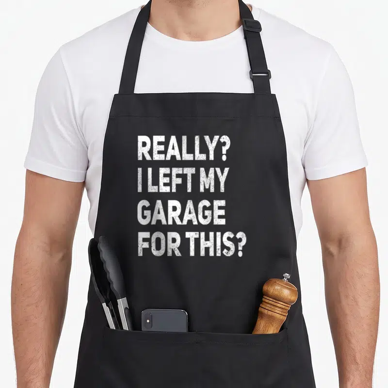 Left garage apron - White stacked text reading Really I Left My Garage For This on black fabric — shown on a black cotton canvas apron against a white background