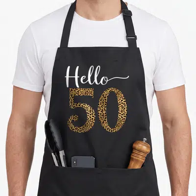 Hello leopard print apron - Hello 50 text with tan and black leopard print pattern filling the numbers — shown on a black cotton canvas apron against a white background