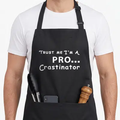 Trust procrastinator apron - White text reading TRUST ME I'M A PRO... Crastinator in three stacked lines — shown on a black cotton canvas apron against a white background