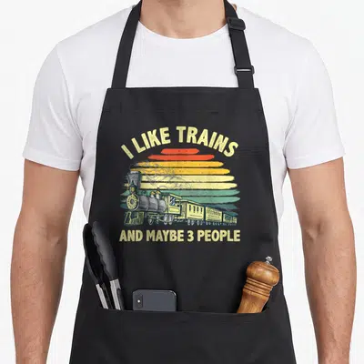 Funny collector railroad train apron - Retro steam train illustration with beige text I LIKE TRAINS AND MAYBE 3 PEOPLE — shown on a black cotton canvas apron against a white background
