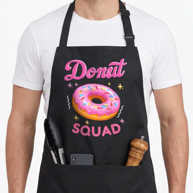 Squad sprinkle apron - Pink frosted donut with sprinkles and text Donut SQUAD surrounded by stars — shown on a black cotton canvas apron against a white background