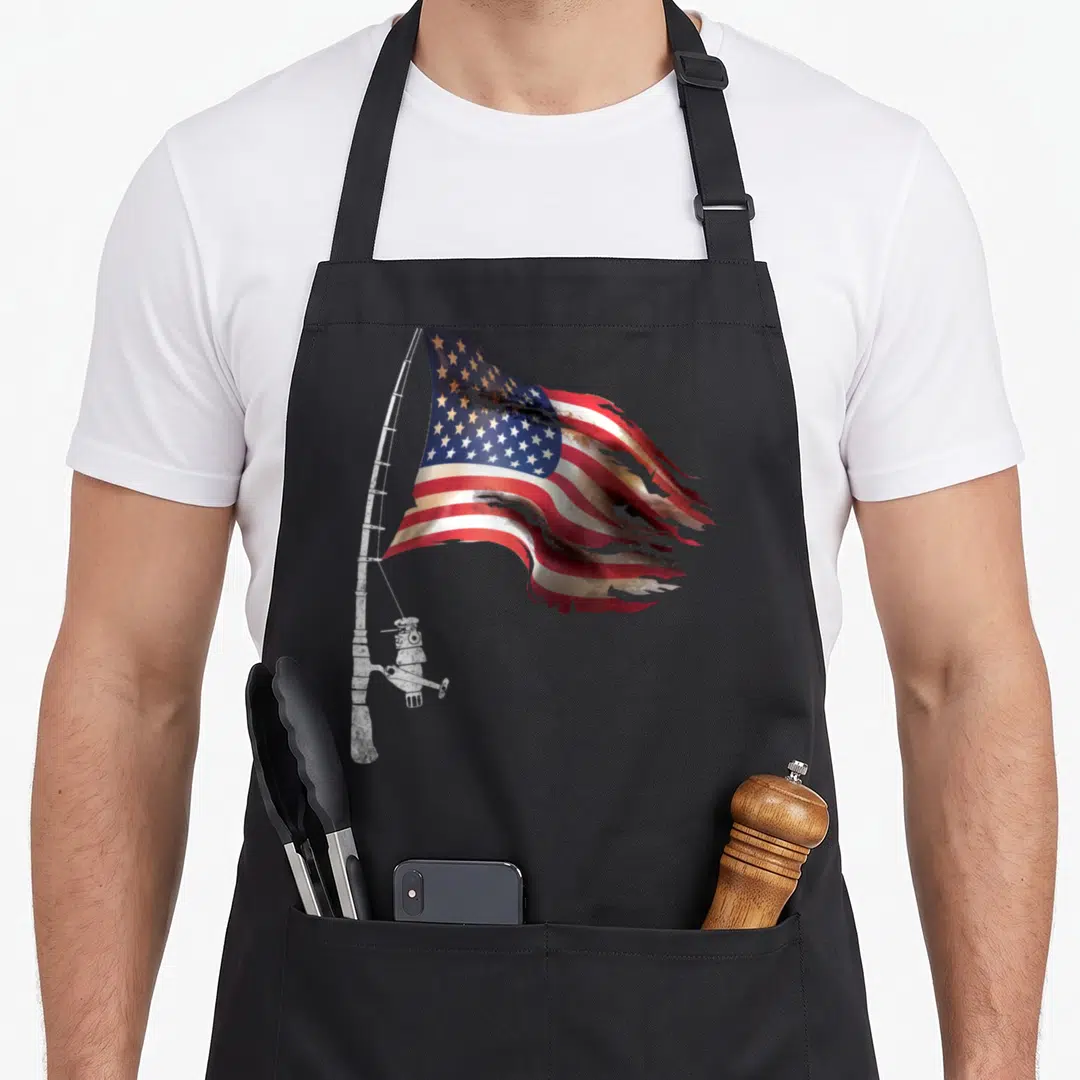 Fisherman 4th of july apron - Tattered USA flag with red white and blue stripes draped over a white fishing rod and reel — shown on a black cotton canvas apron against a white background