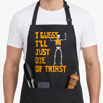 Guess tots apron - Cartoon skeleton in orange vest next to stacked orange text reading I guess I'll just die of thirst — shown on a black cotton canvas apron against a white background