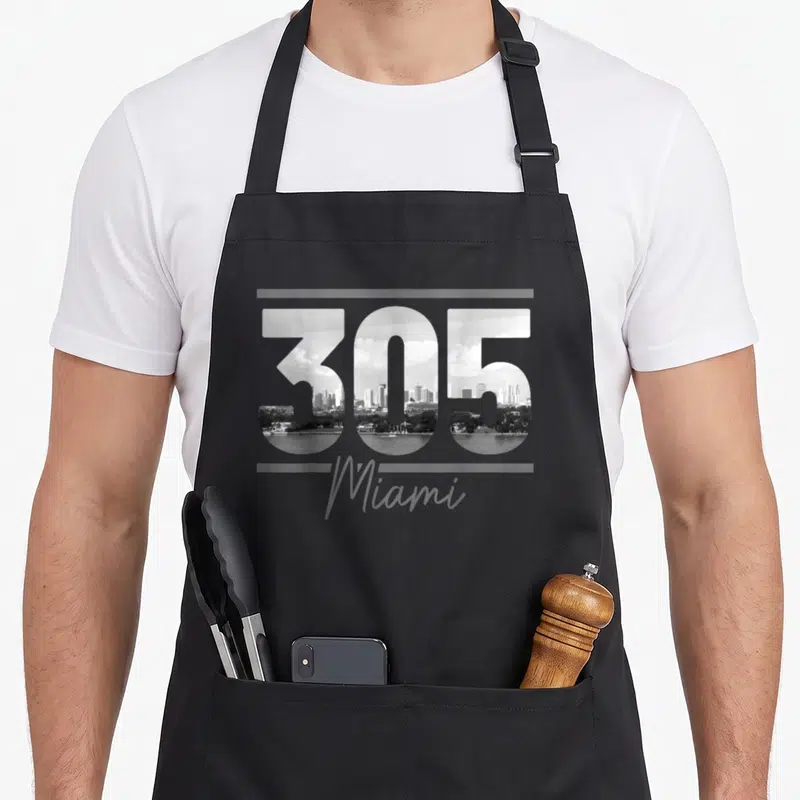 Area florida apron - 305 number filled with cityscape and Miami text in grayscale — shown on a black cotton canvas apron against a white background