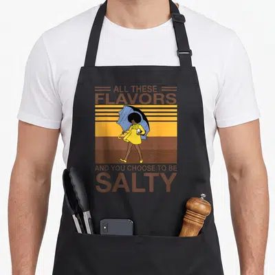 Flavor these apron - Woman in yellow dress holding blue umbrella raining salt with brown text on striped background — shown on a black cotton canvas apron against a white background