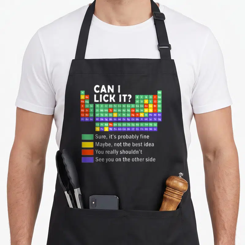 Lick science apron - Periodic table with safety legend reading Can I Lick It and color coded blocks — shown on a black cotton canvas apron against a white background