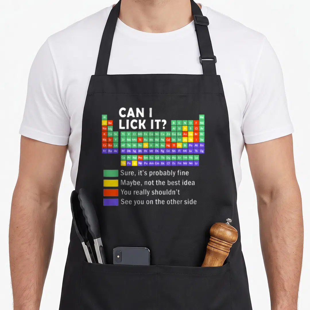 Lick science apron - Periodic table with safety legend reading Can I Lick It and color coded blocks — shown on a black cotton canvas apron against a white background