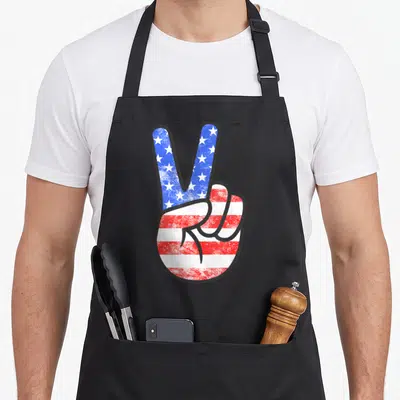 Peace 4th of july apron - Cartoon hand making a peace sign filled with blue stars and red white stripes — shown on a black cotton canvas apron against a white background