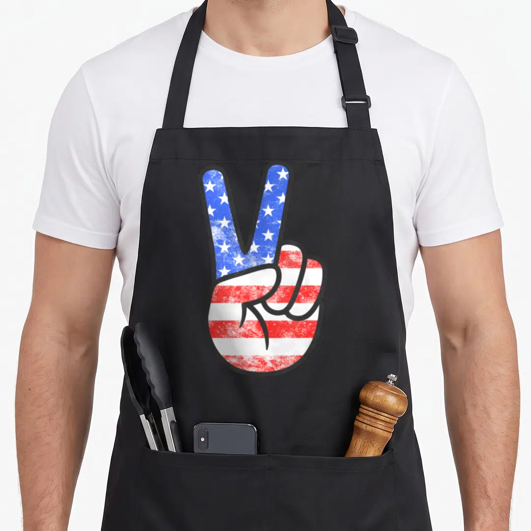 Peace 4th of july apron - Cartoon hand making a peace sign filled with blue stars and red white stripes — shown on a black cotton canvas apron against a white background