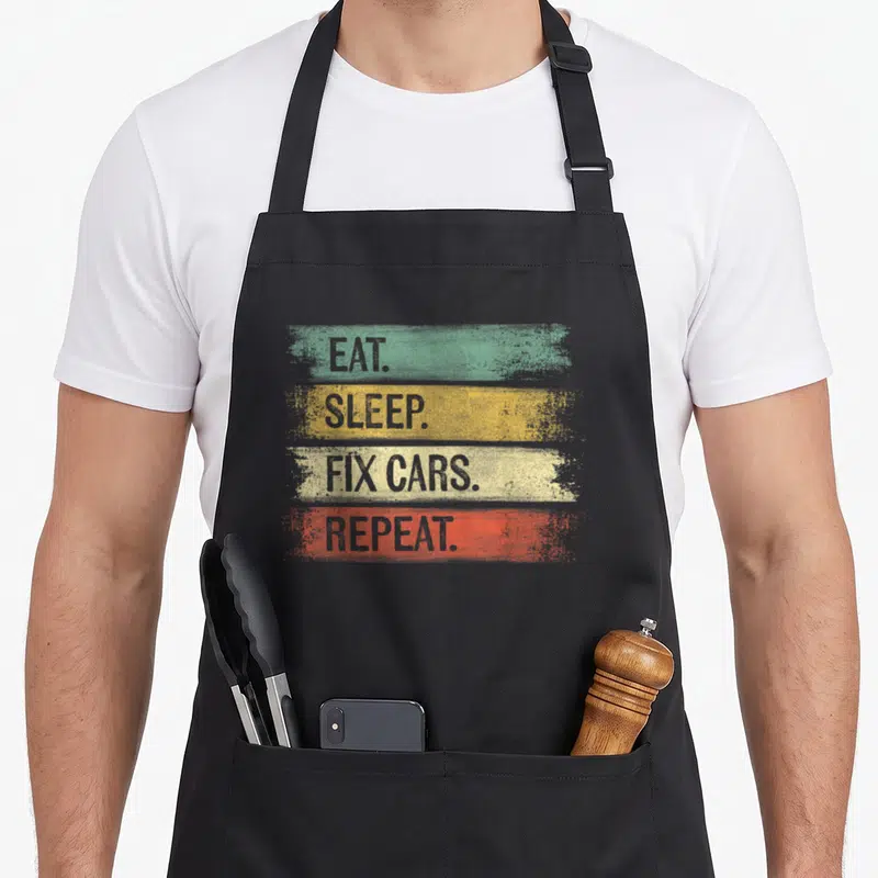 Eat auto apron - Stacked text Eat Sleep Fix Cars Repeat on green yellow beige and orange brushstrokes — shown on a black cotton canvas apron against a white background