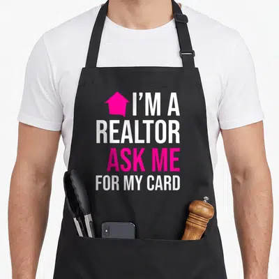 Ask real estate agent apron - Pink house icon next to stacked white and pink text reading I'M A REALTOR ASK ME FOR MY — shown on a black cotton canvas apron against a white background