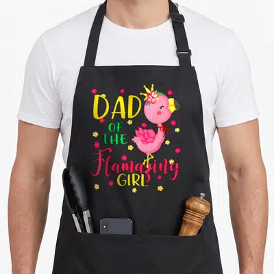 Flamazing flamingo apron - Pink flamingo wearing a crown next to stacked yellow and red text reading Dad — shown on a black cotton canvas apron against a white background