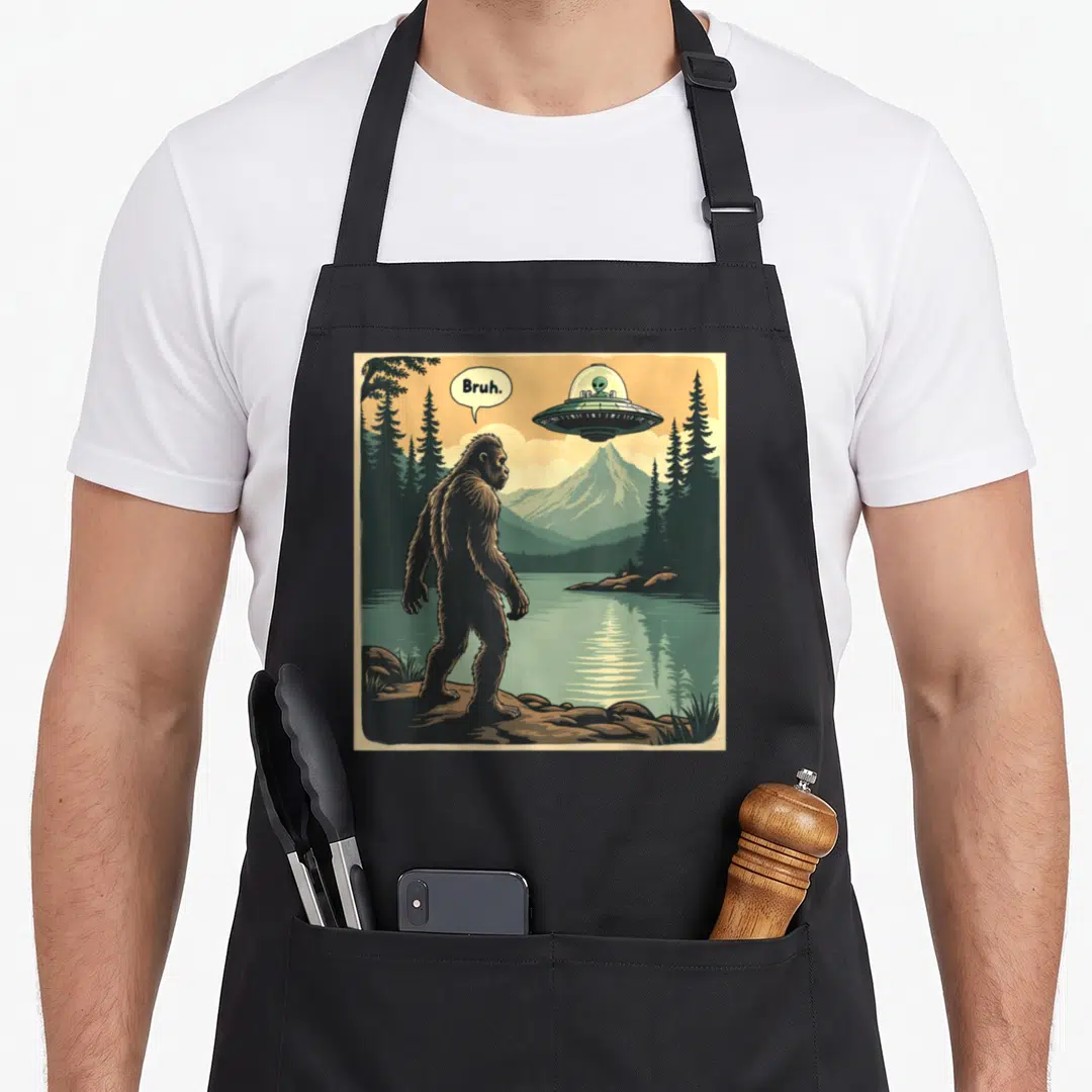 Spaceship bigfoot apron - Cartoon Bigfoot saying Bruh to a UFO in a forest scene with mountains and lake — shown on a black cotton canvas apron against a white background