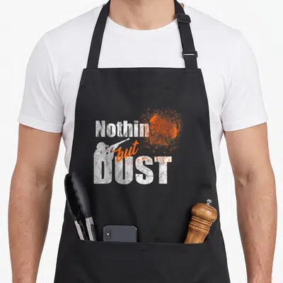 Trap clay apron - Nothin but DUST text with white shooter silhouette and orange exploding clay graphic — shown on a black cotton canvas apron against a white background