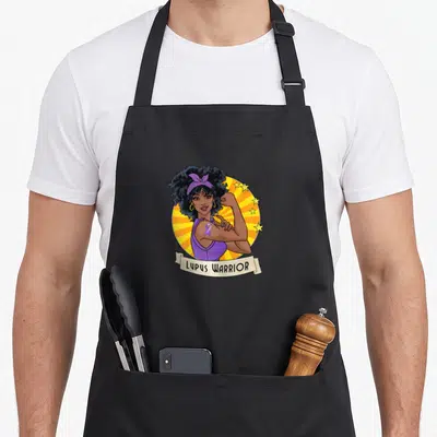 Funny warrior lupus awareness apron - Cartoon woman flexing arm with purple ribbon and LUPUS WARRIOR text on beige — shown on a black cotton canvas apron against a white background