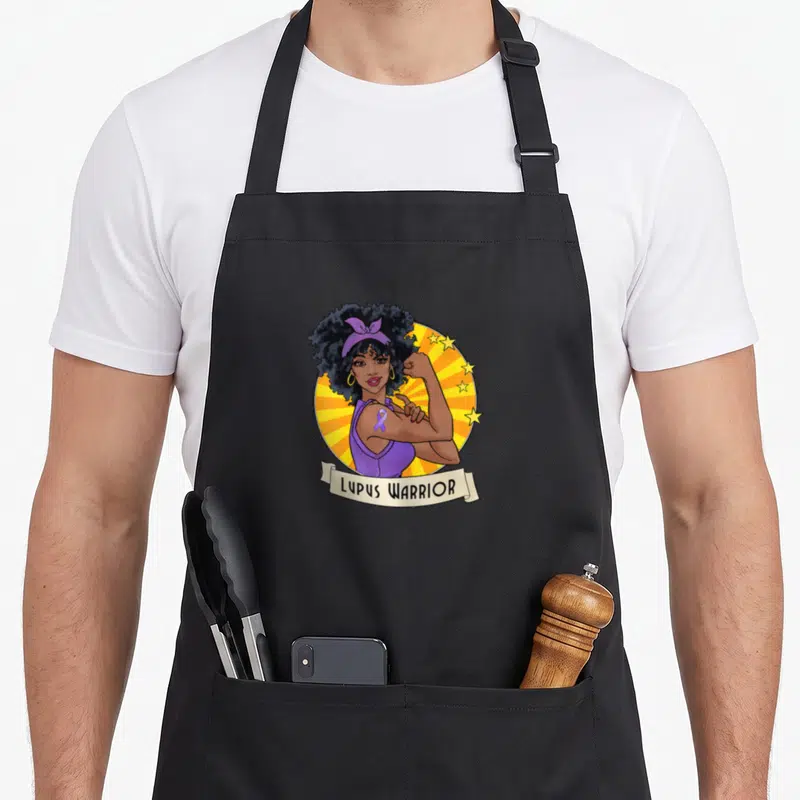 Funny warrior lupus awareness apron - Cartoon woman flexing arm with purple ribbon and LUPUS WARRIOR text on beige — shown on a black cotton canvas apron against a white background