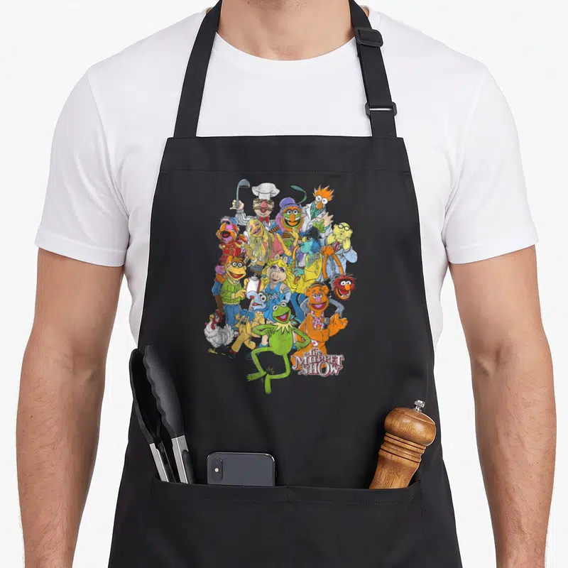 Main character aprons - Kermit the Frog at bottom center of dense Muppet cluster with show title to right — shown on a black cotton canvas apron against a white background
