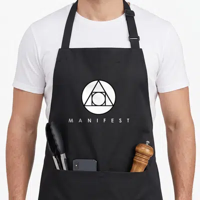 Symbol manifestation apron - Black circle triangle symbol above white MANIFEST text on grey background — shown on a black cotton canvas apron against a white background