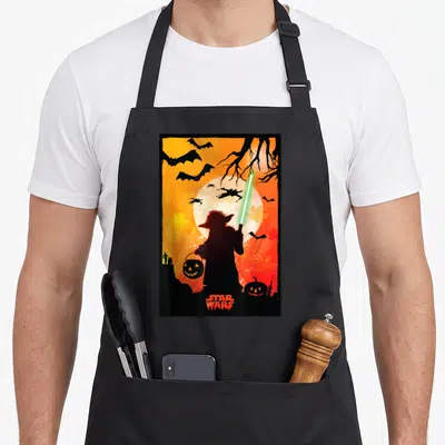 Yoda halloween apron - Yoda silhouette holding green lightsaber and pumpkin bucket against orange moon with TIE fighters — shown on a black cotton canvas apron against a white background
