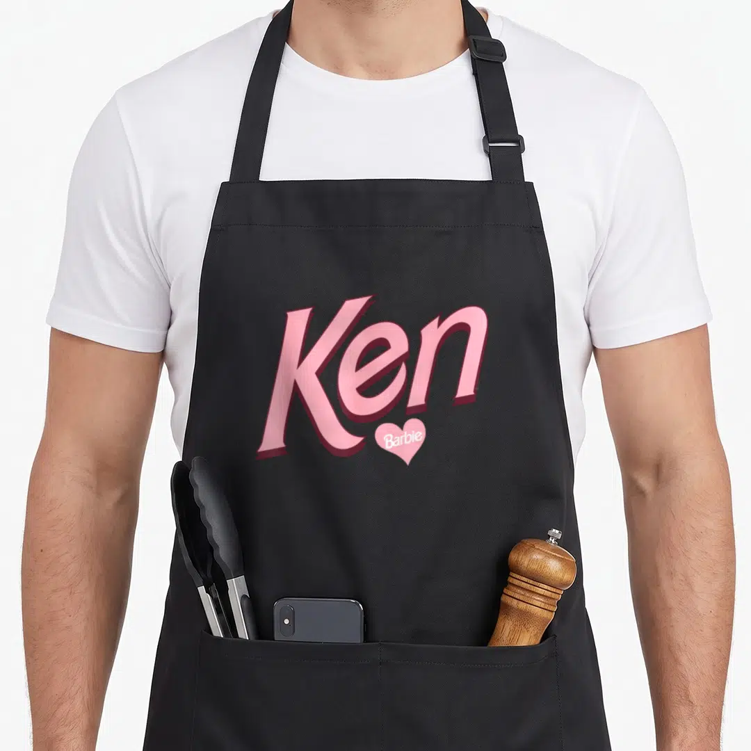 Funny ken general apron - Pink Ken text with small white Barbie word inside a pink heart below the e — shown on a black cotton canvas apron against a white background