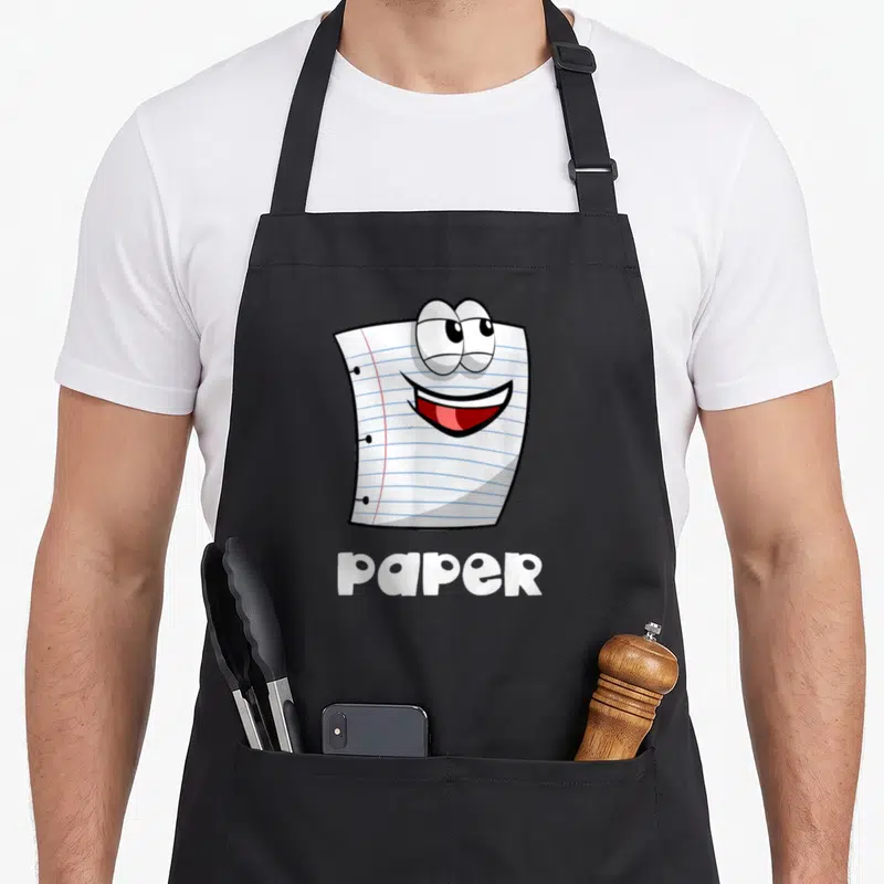 Scissor halloween paper apron - Cartoon white lined paper sheet with face and bold paper text — shown on a black cotton canvas apron against a white background