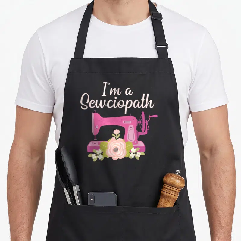 Sewing grandma apron - Pink sewing machine with I'm a Sewciopath text and floral cluster at base — shown on a black cotton canvas apron against a white background