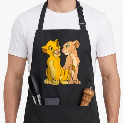 Together lion apron - Two cartoon lion cubs sitting side by side in yellow and tan with black outlines — shown on a black cotton canvas apron against a white background