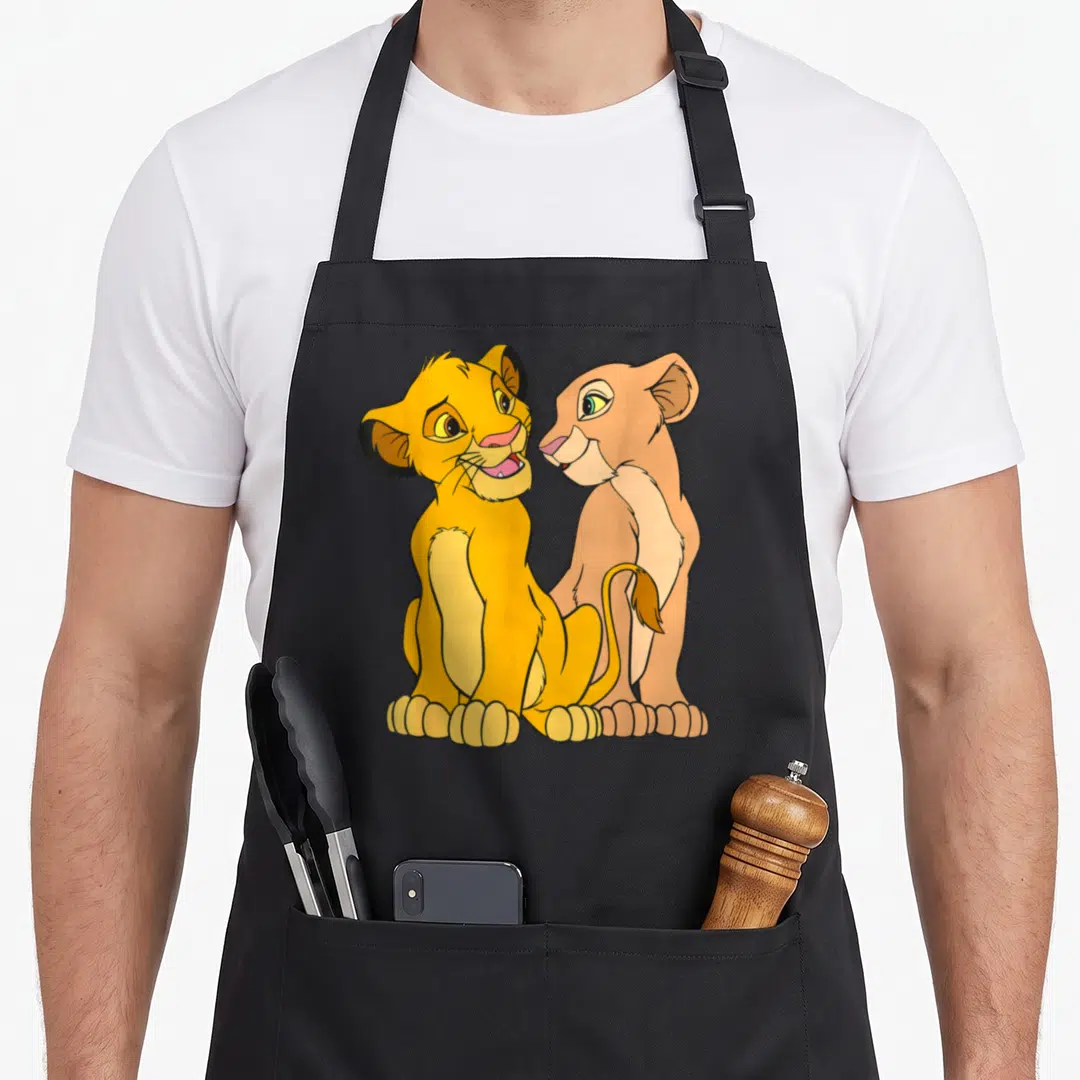 Together lion apron - Two cartoon lion cubs sitting side by side in yellow and tan with black outlines — shown on a black cotton canvas apron against a white background