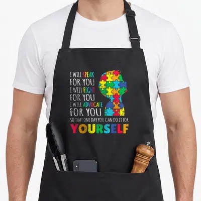 Support mom and son aprons - Stacked advocate text next to a head silhouette filled with multicolored puzzle pieces — shown on a black cotton canvas apron against a white background