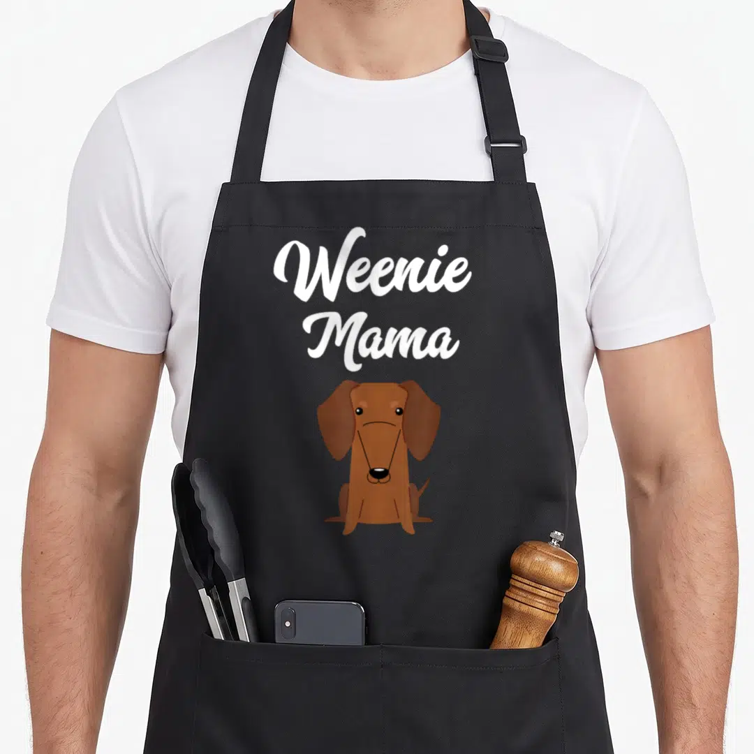 Mama dachshund apron - Weenie Mama text above sitting brown cartoon dachshund on black background — shown on a black cotton canvas apron against a white background