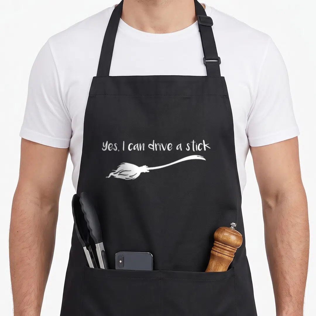 Stick halloween witch apron - White yes I can drive a stick text above a horizontal broomstick graphic — shown on a black cotton canvas apron against a white background