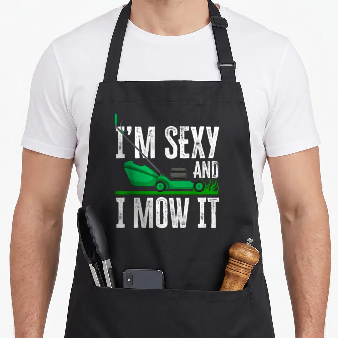 Mow gardener apron - Green lawn mower on grass strip with I'M SEXY AND I MOW IT text — shown on a black cotton canvas apron against a white background