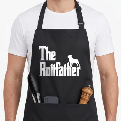 Rottfather rottweiler apron - White stacked text The Rottfather with a standing dog silhouette on black fabric — shown on a black cotton canvas apron against a white background