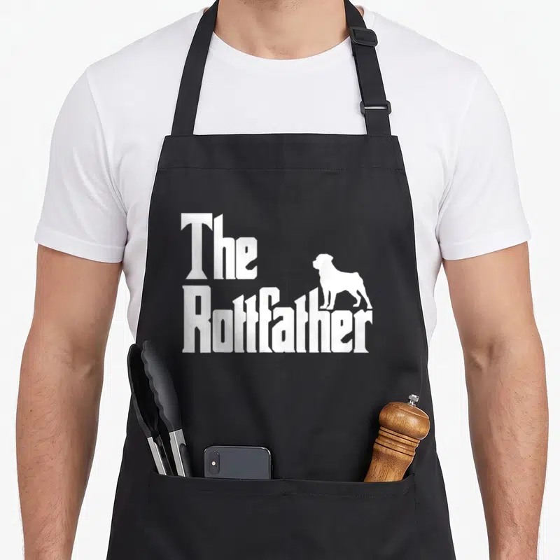 Rottfather rottweiler apron - White stacked text The Rottfather with a standing dog silhouette on black fabric — shown on a black cotton canvas apron against a white background