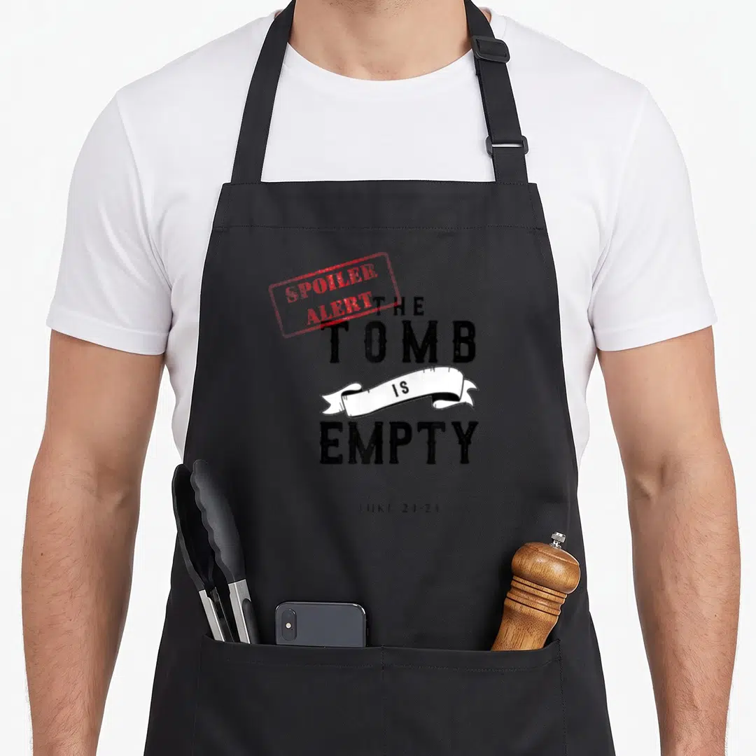 Christian spoiler easter apron - Red spoiler alert stamp over black The Tomb Is Empty text with white ribbon and Luke — shown on a black cotton canvas apron against a white background