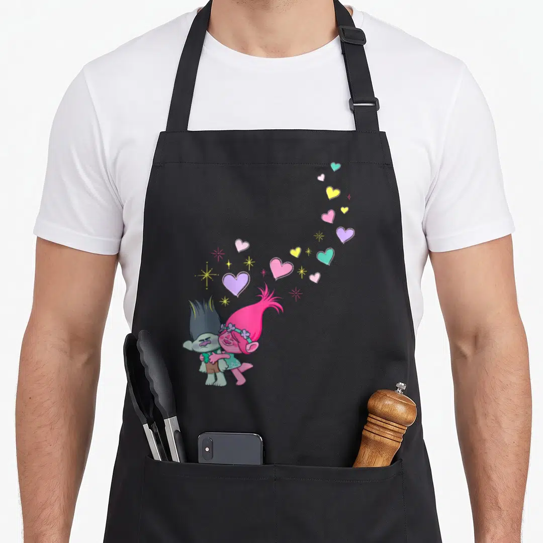 Cute troll apron - Two cartoon trolls hugging with a trail of pink, purple, yellow, and teal hearts and stars curving — shown on a black cotton canvas apron against a white background