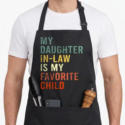 Family mom and daughter aprons - Stacked colorful text reading MY DAUGHTER IN-LAW IS MY FAVORITE CHILD in green yellow — shown on a black cotton canvas apron against a white background