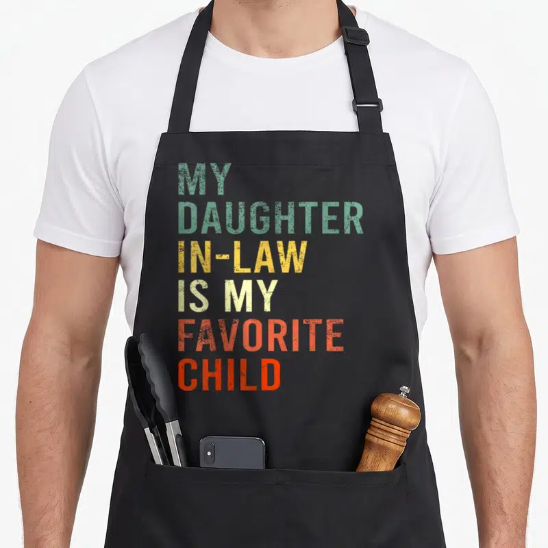 Family mom and daughter aprons - Stacked colorful text reading MY DAUGHTER IN-LAW IS MY FAVORITE CHILD in green yellow — shown on a black cotton canvas apron against a white background